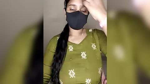 Media: Video of a South Asian woman with a long braid, wearing a black mask and olive-green top with floral patterns, covering her face and chest. Background is blurred.