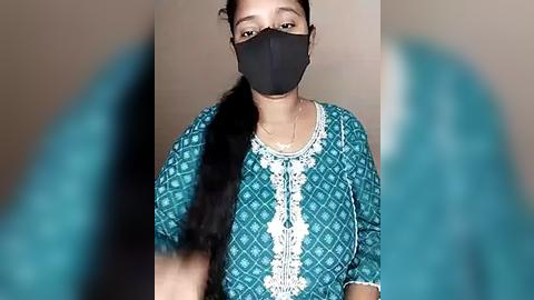 Media: A video of a South Asian woman with medium skin tone, wearing a teal patterned sari blouse, black face mask, and black hair in a ponytail, standing against a beige wall.