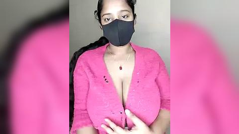 Media: Video of a light-skinned woman with large breasts, wearing a pink cardigan, black face mask, and dark hair in a ponytail, standing against a plain, light-colored wall.