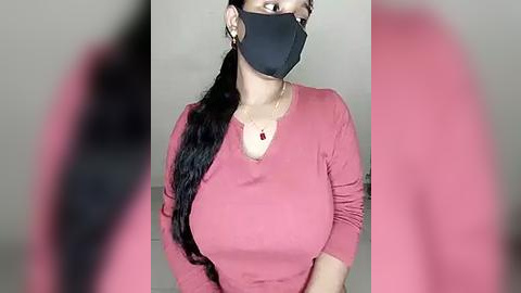 Media: A video of a woman with medium skin tone, wearing a black face mask, black earrings, and a pink long-sleeve top, with long black hair in a ponytail, standing indoors against a blurred background.