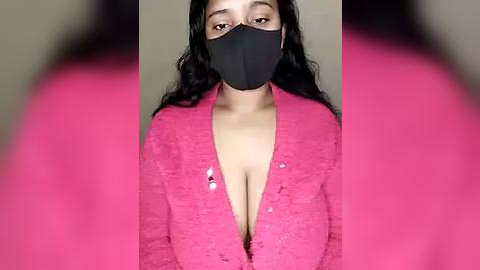 Media: Video of a young woman with long black hair, wearing a pink lace cardigan that reveals her cleavage, a black face mask, and a blurred background.