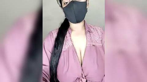 Media: Video of a woman with a black face mask, wearing a pink blouse with lace detailing, her long black hair tied in a ponytail, blurred background.