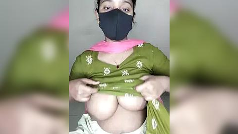 Media: Video of a woman with a dark complexion, wearing a green shirt with white floral patterns, exposing large, bare breasts. She has a pink scarf around her neck, a black mask covering her nose and mouth, and is indoors.