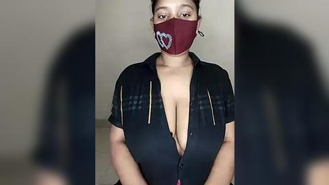 Media: A video of a plus-sized woman with medium skin tone, wearing a maroon face mask and a low-cut black blouse, standing in a plain, indoor setting.