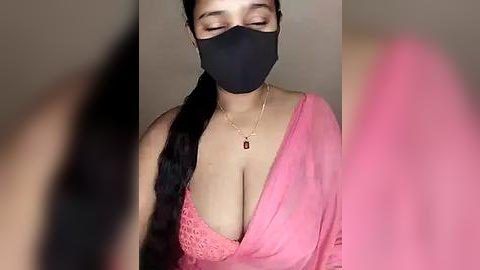 Media: A video of a South Asian woman with medium skin tone and long black hair, wearing a black face mask and a low-cut pink saree, revealing ample cleavage. She has a delicate gold necklace with a small pendant. The background is blurred.