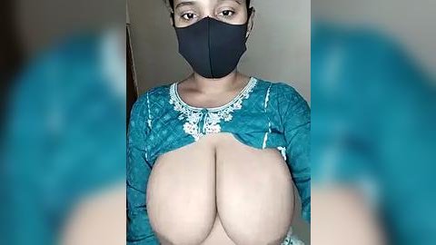 Media: A video of a woman with dark skin, wearing a teal embroidered blouse, black face mask, and blue hair extensions, exposing her large, natural breasts.