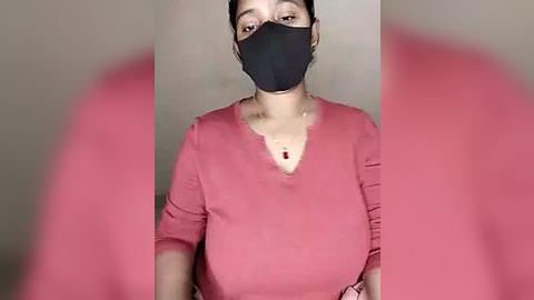 Media: A video of a young woman with fair skin, black hair pulled back, wearing a black face mask and a pink V-neck top, standing against a neutral background.