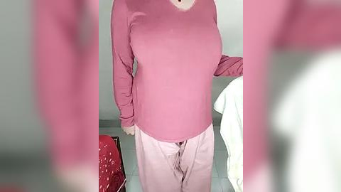 Media: Video of a person wearing a pink sweater and white pants, standing in a room with pale walls and a red chair.