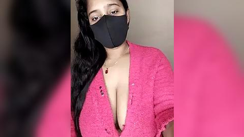 Media: Video of a light-skinned woman with long black hair wearing a black face mask, pink lace cardigan, and black bra, showing cleavage. Blurry background.