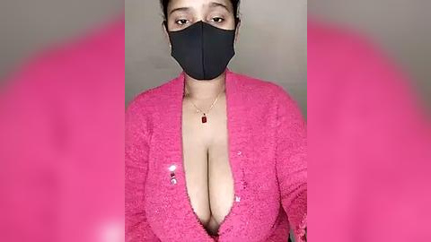 Media: A video of a woman with medium skin tone and a full bust, wearing a pink lace cardigan with a plunging neckline, black face mask, and red pendant necklace.