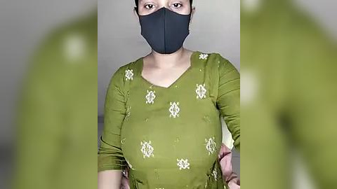 Media: A video of a woman with a medium complexion, wearing a black face mask, green top with white floral embroidery, and pink shawl, standing indoors with blurred green background.