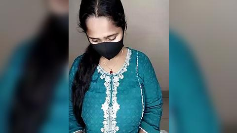 Media: Video of a South Asian woman with medium skin tone, wearing a teal long-sleeved kurta with intricate embroidery, black face mask, and black hijab, kneeling on a wooden floor, blurred background.