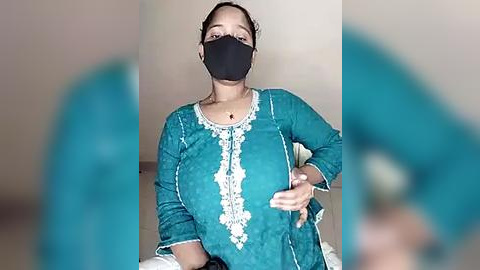 Media: Video of a South Asian woman with medium skin tone and black hair tied back, wearing a teal kurta with white embroidery, black mask, and seated indoors.