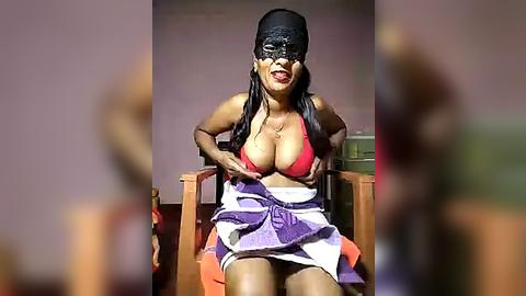 Media: Video of a woman with medium-dark skin, wearing a black mask, red bra, and purple skirt, sitting in a wooden chair.