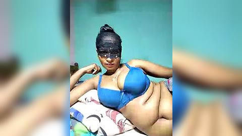 Media: Video of a plus-size woman with medium brown skin, wearing a blue bikini top, lying on a colorful beach towel, with a mask covering her eyes.