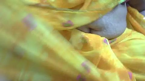 Media: A close-up video of a person's bare torso wrapped in a vibrant yellow sari with subtle pink floral patterns. The sari's fabric is draped over the person's chest, partially covering a light-skinned, smooth abdomen.