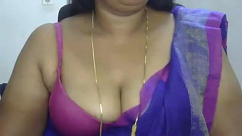 Media: A close-up video of a woman with medium skin tone wearing a purple and pink bra, draped in a blue and purple sari, with a gold necklace.