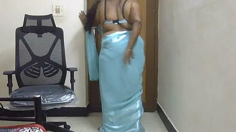 Media: A video shows a dark-skinned woman with long hair in a light blue satin sari, standing in a small, dimly lit room with a black ergonomic chair and a wooden door in the background.