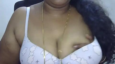 Media: Video of a woman with medium brown skin, wearing a white bra with cloud patterns and a gold chain necklace. Her full, round breasts are prominent. Background is out of focus.