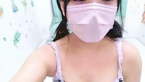 Media: Video of an Asian woman with long black hair, wearing a light pink surgical mask, a white lace camisole, and teal earrings, taken from a slightly elevated angle. Background features a white wall with green floral decals.