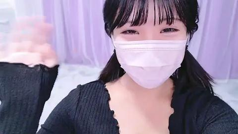 Media: Video of a young East Asian woman with straight black hair and bangs, wearing a black ribbed sweater and a white face mask, in a softly lit room with purple curtains.