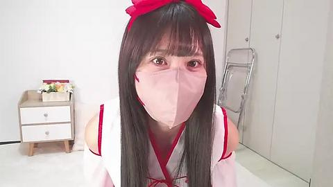 Media: Video of an Asian woman with long black hair and a red ribbon headband, wearing a white and red kimono with a face mask, in a white room with a wooden cabinet, dresser, and flower vase.