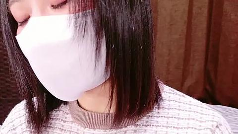 Media: Close-up video of an Asian woman with shoulder-length, straight black hair, wearing a white surgical mask, a beige ribbed sweater, and a dark brown textured background.