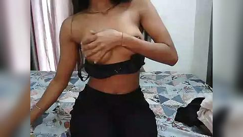 Media: Video of a slender, light-skinned woman with long, straight black hair, topless, covering her breasts with her hands, wearing high-waisted black pants. She is in a pastel-colored bedroom with a quilted bedspread.