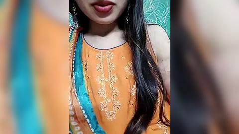 Media: A video of a woman with long, dark hair, wearing an orange kurta with intricate embroidery and a teal dupatta. She has light brown skin and is partially in focus, with a blurred background.