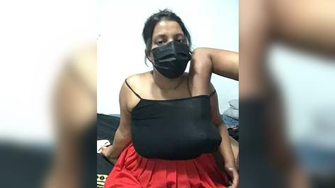 Media: Video of a plus-size woman with medium brown skin and dark hair, wearing a black mask, black tank top, and red pleated skirt, sitting on a bed, blurred figures in the foreground.