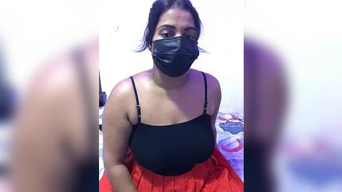 Media: Video of a woman with dark skin and black hair, wearing a black face mask, black spaghetti-strap top, and red pleated skirt, sitting on a bed with a white wall background.