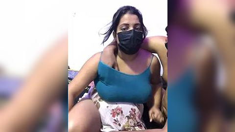 Media: Video of a woman with medium brown skin and long black hair wearing a teal tank top, floral skirt, black mask, and sitting on a bed in a dimly lit room with blurred figures in the background.