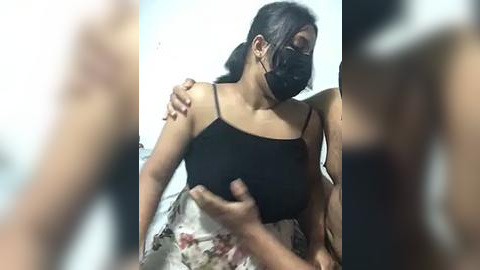 Media: Video of a woman with medium skin tone, dark hair, wearing a black face mask, black spaghetti strap tank top, and floral skirt. Her breasts are being grabbed from behind by an unseen person. The background is blurred, with the woman's expression hidden by the mask.