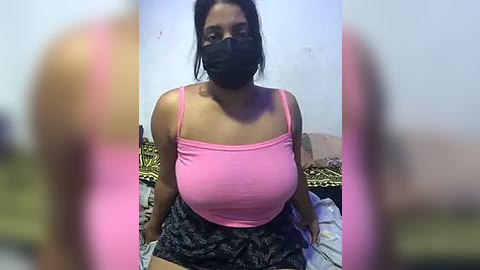 Media: Video of a curvy woman with a medium complexion, wearing a pink spaghetti-strap top and black shorts with a floral pattern. She has dark hair, a black face mask, and sits on a bed with a leopard-print blanket.