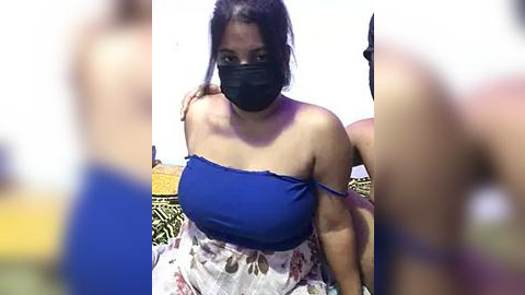 Media: Video of a woman with medium brown skin, wearing a blue top and floral skirt, black mask, and casual attire, standing outdoors against a blurred background.