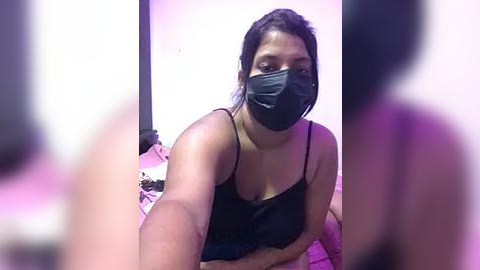 Media: Video of a dark-skinned woman with a medium build, wearing a black face mask, black lace bra, and black tank top, sitting on a bed in a dimly lit room with purple lighting.