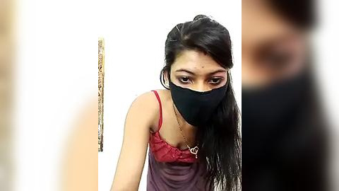 Media: Video of a young woman with long black hair, wearing a red spaghetti-strap top, black face mask, and a gold necklace, standing against a white background, holding a beige cane.