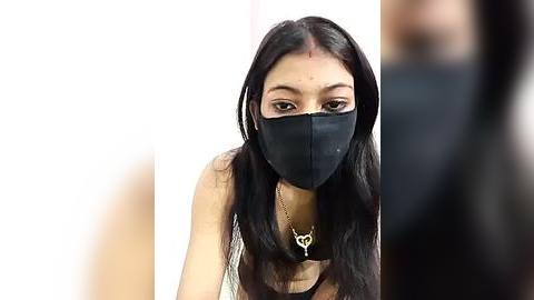 Media: Video of a woman with long black hair, wearing a black mask and a silver necklace, standing against a plain white background.