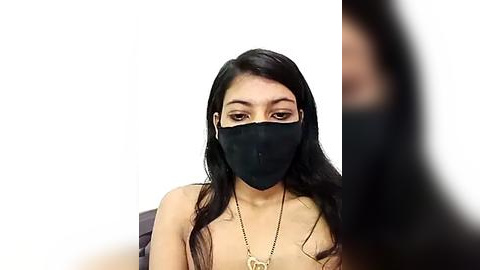 Media: Video of a young, light-skinned Asian woman with long black hair, wearing a black mask and necklace, seated indoors against a plain white background.