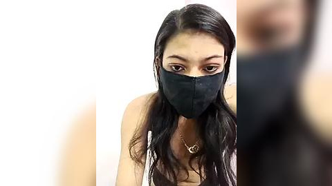 Media: Video of a young Asian woman with long black hair, wearing a black face mask, a white top, and a gold necklace, against a plain white background.