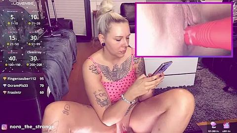 Media: A blonde woman with tattoos, wearing a pink polka-dot top, sits on a couch, holding a phone, while a close-up shows a pink vibrator inserted into her vagina.