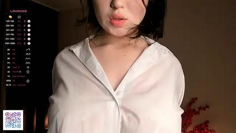 Media: Video of a young Asian woman with pale skin, wearing a white button-up shirt, partially unbuttoned, revealing her cleavage. Background shows dimly lit room with a digital clock and red flowers.