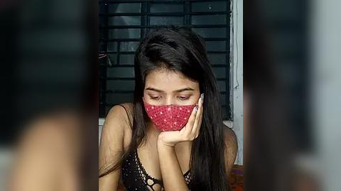 Media: Video of a young woman with long black hair, wearing a red facemask and black lace top, standing indoors against a dark, barred window background.