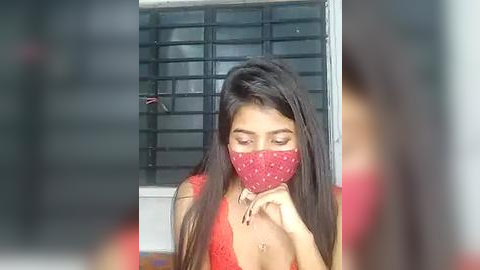 Media: Video of a young woman with long, straight black hair, wearing a red mask and red lace top, standing indoors with blurred background.