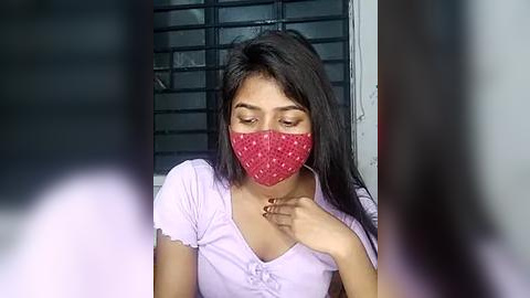 Media: Video of a young South Asian woman with long dark hair, wearing a red patterned face mask, white short-sleeved top, and resting hand on chest. Background shows a barred window and darkened room.