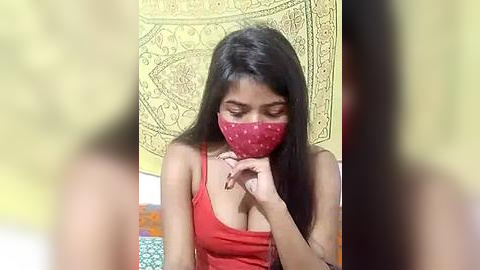 Media: Video of a young South Asian woman with long black hair, wearing a red mask, tank top, and sitting indoors against a yellow tapestry backdrop.