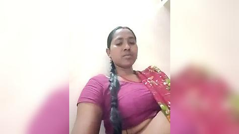 Media: Video of a woman with medium-dark skin, long braided hair, wearing a pink crop top and a red sari with floral patterns. Background shows a plain beige wall.