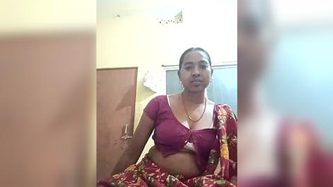 Media: A video of an Indian woman with medium skin tone and dark hair, wearing a red and yellow floral sari, seated indoors with a wooden door and white wall background.