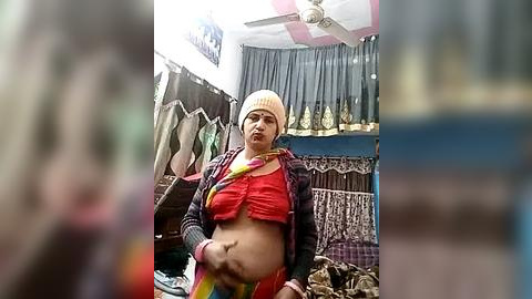 Media: Video of a pregnant woman in a colorful, patterned saree and beige beanie, standing in a crowded, cluttered indoor market with hanging textiles and decorative items.