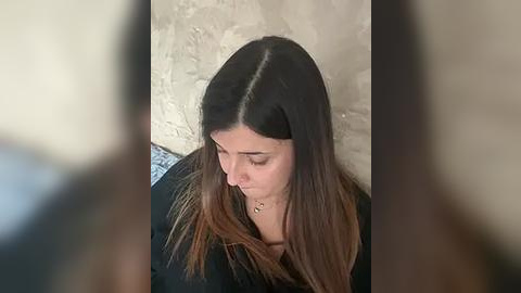 Media: Video of a young woman with straight, dark hair and light brown tips, wearing a black top, looking down thoughtfully. She sits against a textured, beige wall. Background is slightly blurred.